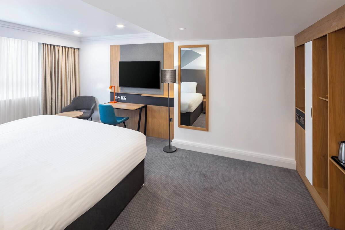 A hotel room with a large bed, a desk with a chair, a flat-screen TV, a mirror, and a wardrobe.