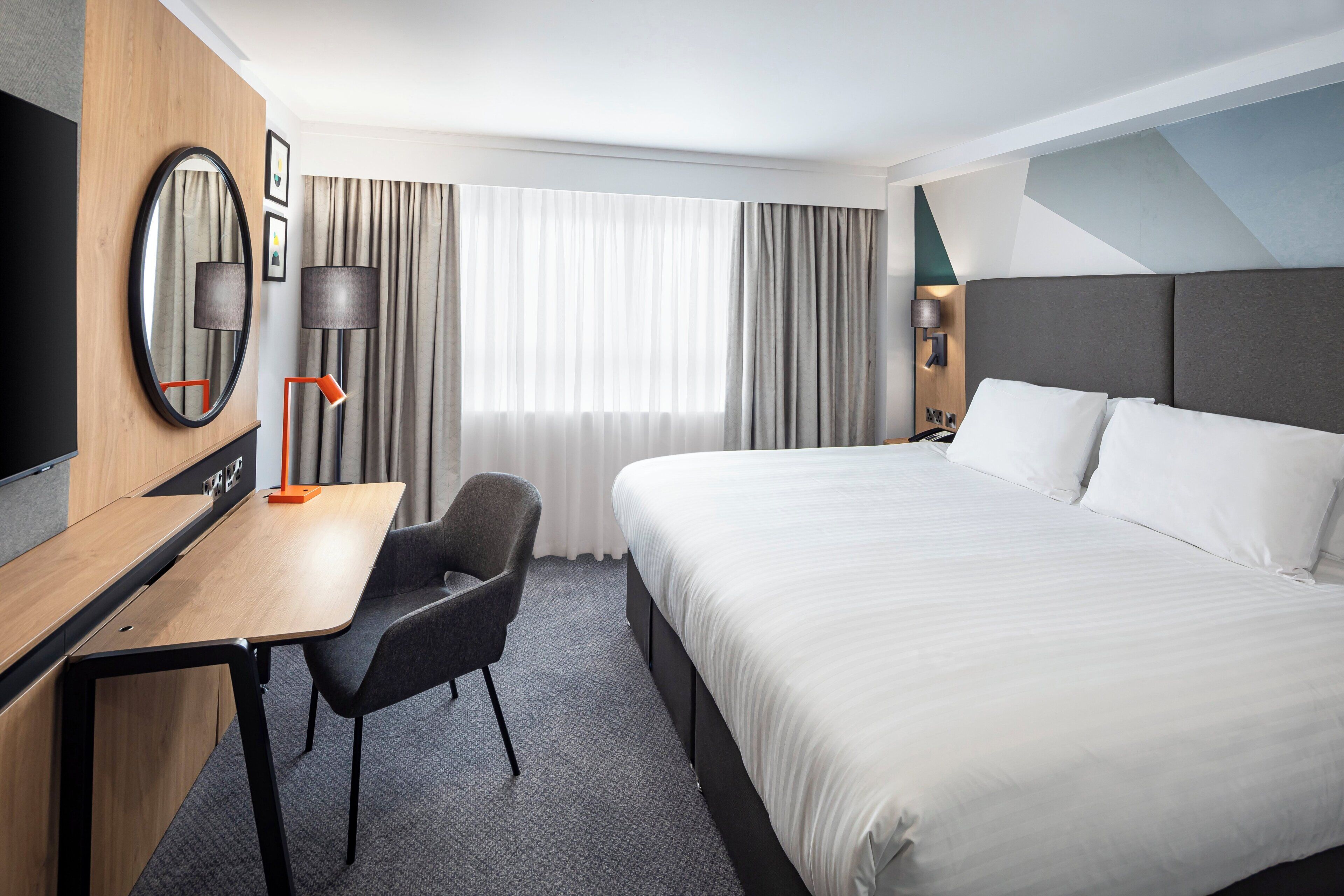 A hotel room with a bed, a desk, a chair, a TV, and a window with curtains.