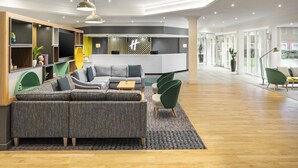 A modern living room with a sectional sofa, a coffee table, and a TV. There is a reception desk with a logo and a plant. The room has wooden flooring and a large window. - Holiday Inn Aylesbury by IHG (Aylesbury)