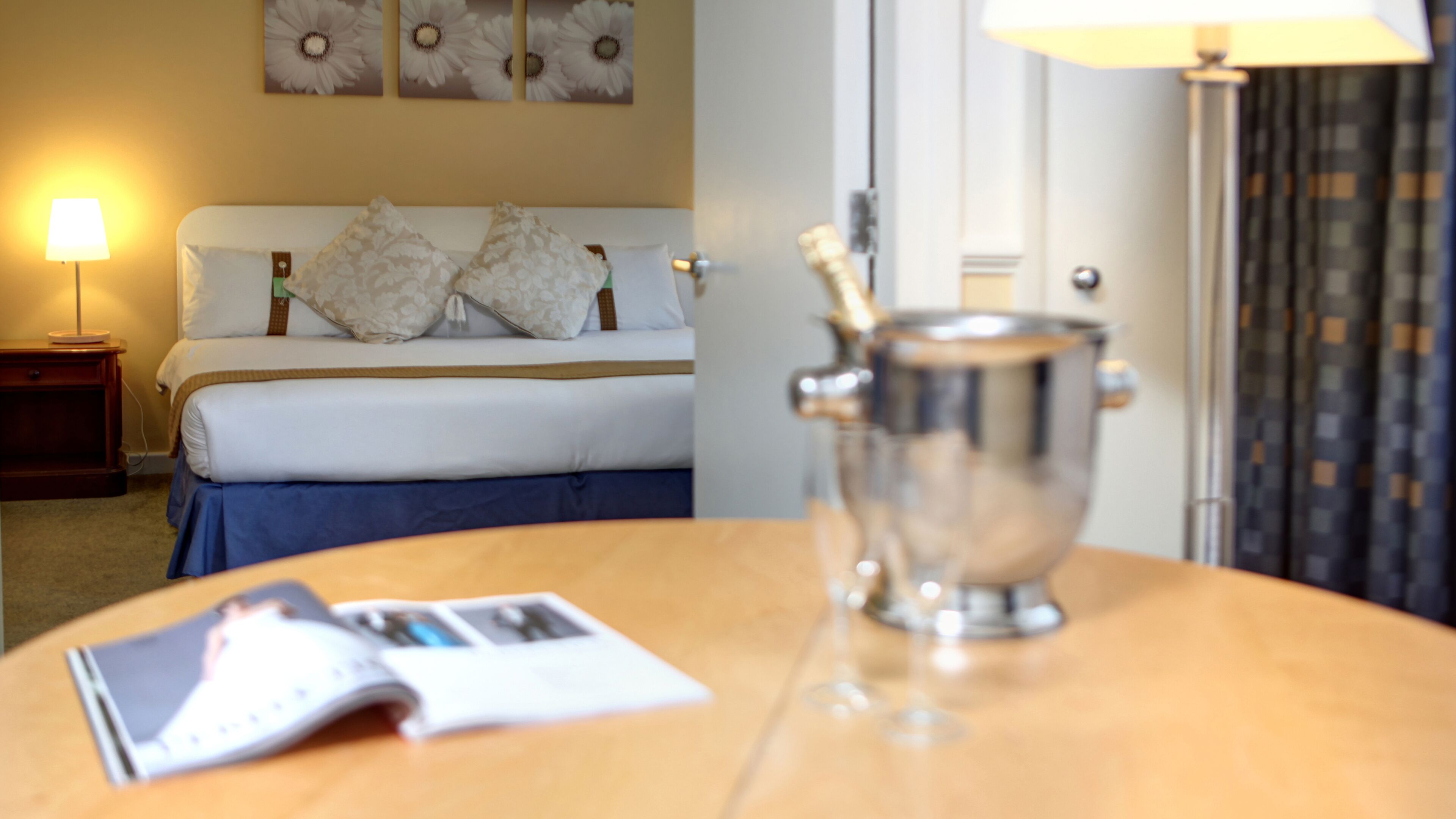 A hotel room with a bed, a nightstand, a lamp, a magazine, and a minibar.