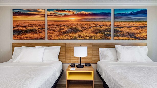 A hotel room with two beds, a wooden headboard, a nightstand with a lamp, and three wall-mounted sunset landscape paintings.