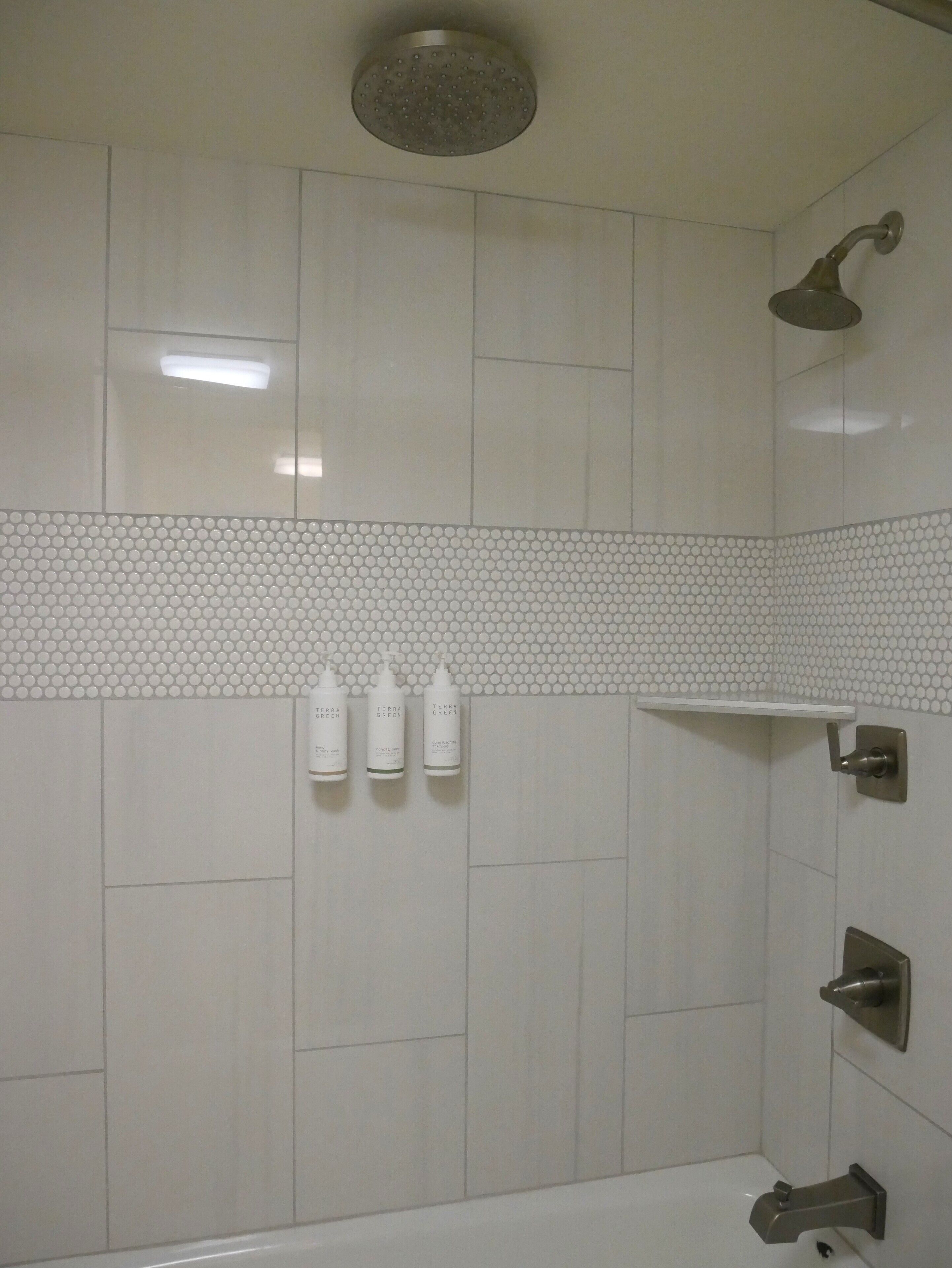 Een badkamer met witte tegels, een douchekop, een badkuip en drie flessen Terra Green shampoo.