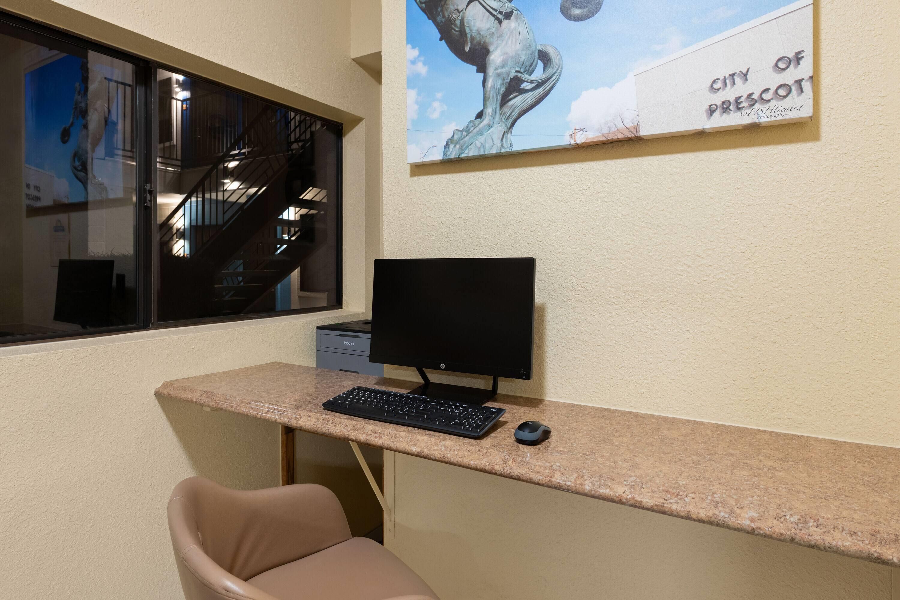 Escritorio de computadora con monitor, teclado y ratón. Una silla se encuentra frente al escritorio. Se ve una ventana con vistas a una escalera y una estatua.