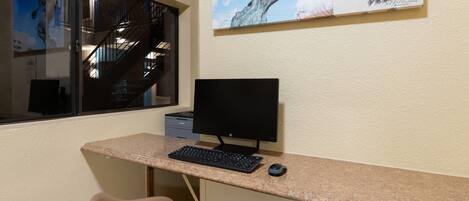 Uma secretária com um monitor, teclado e rato. Uma cadeira está colocada à frente da secretária. Uma janela mostra uma vista de uma escadaria e uma estátua.