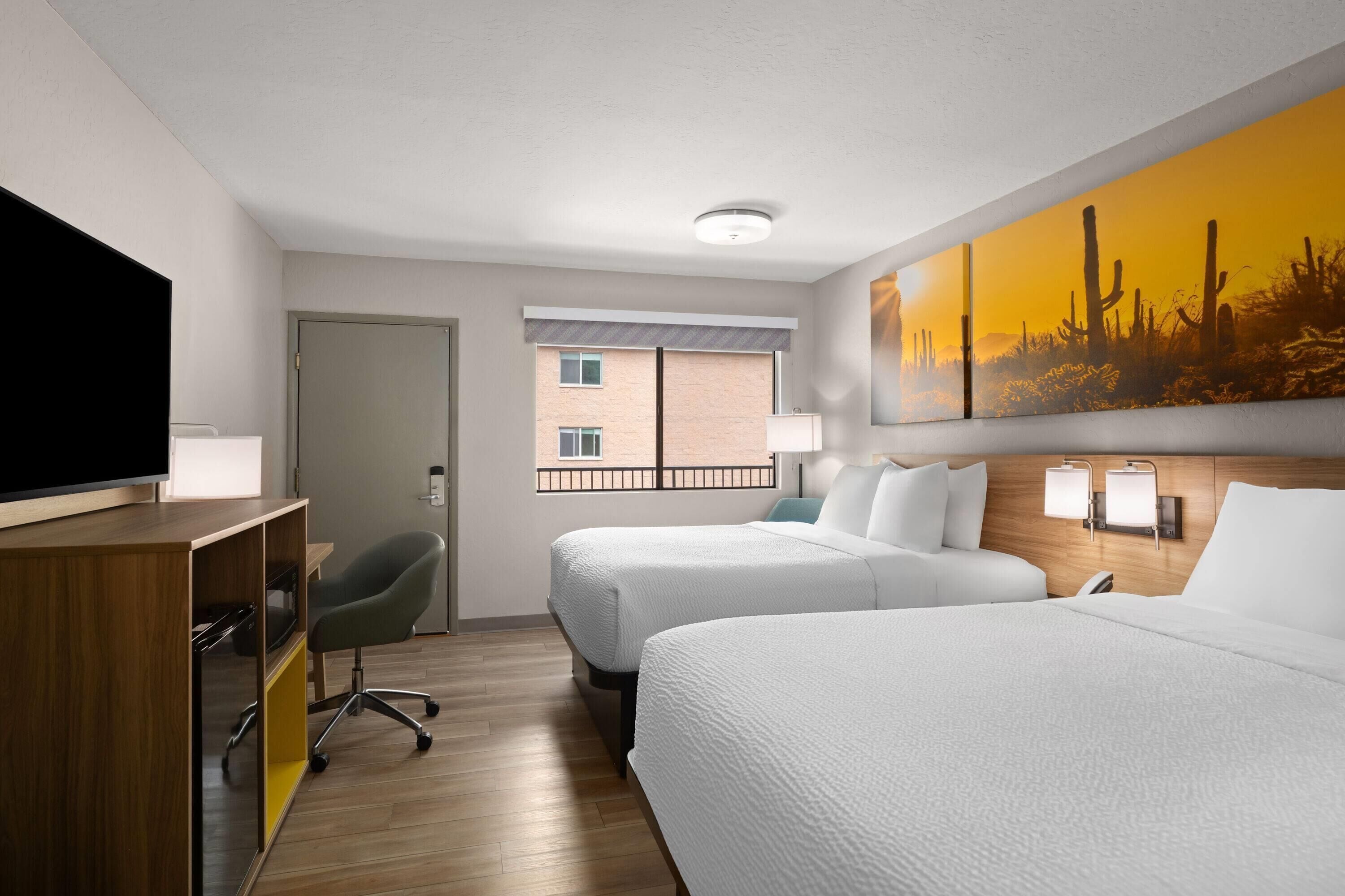 A hotel room with two beds, a TV, a desk, and a painting of a desert landscape.