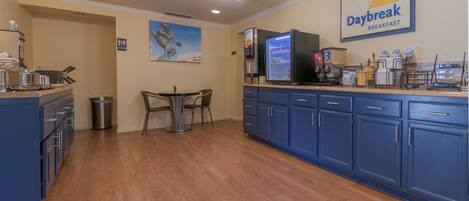 A kitchen with a refrigerator, microwave, and coffee maker. There is a dining table with chairs and a trash can. The kitchen has blue cabinets and a sign for Daybreak Breakfast.