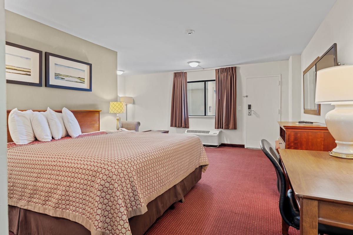 Une chambre d’hôtel avec un grand lit, un bureau, une chaise, une lampe, une fenêtre avec des rideaux et des tableaux encadrés accrochés au mur.