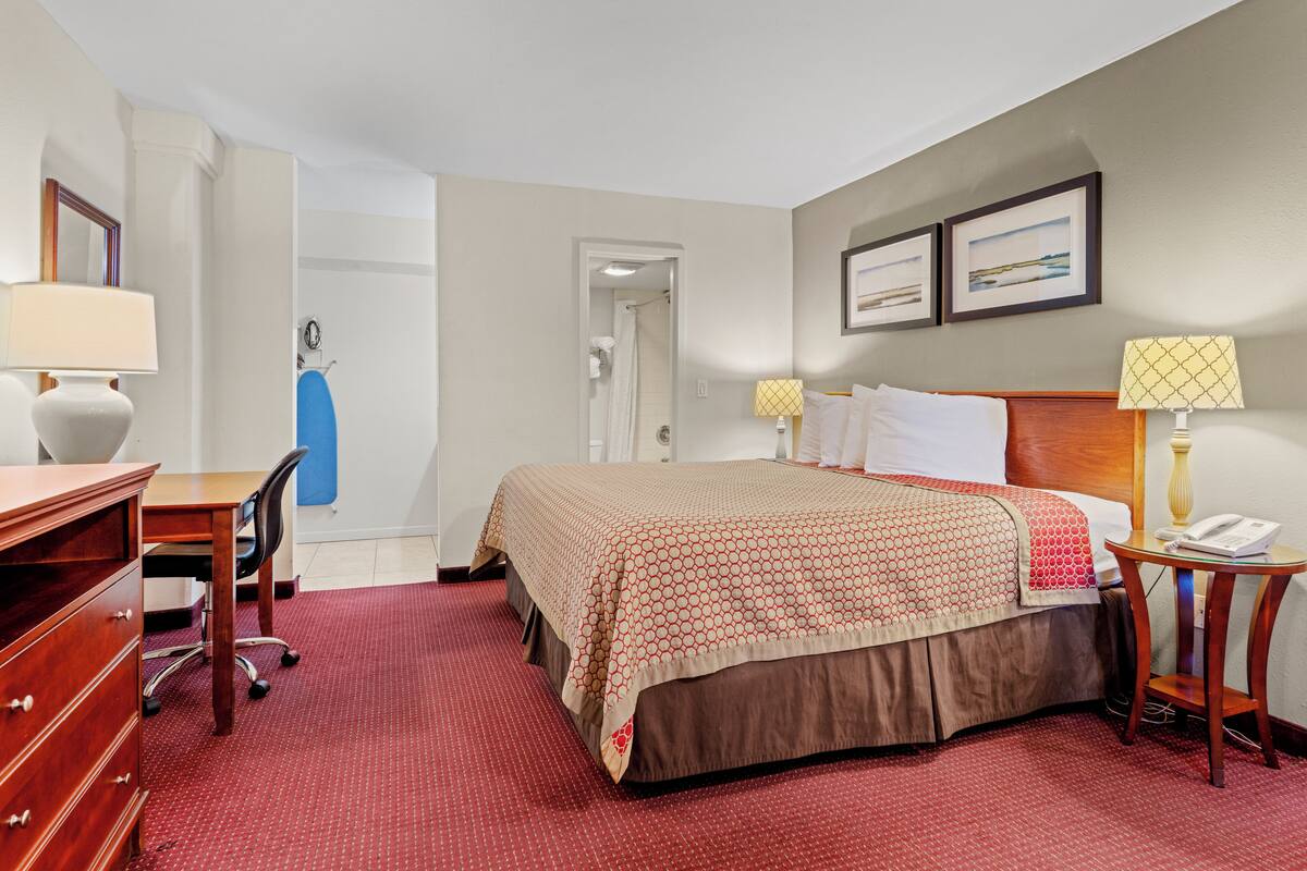 Une chambre d’hôtel avec un lit, un bureau, une chaise, une commode et une salle de bain visible à travers une porte ouverte.