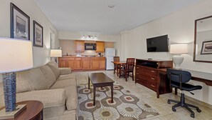 A living room with a sofa, coffee table, and TV. There is a kitchen in the background with wooden cabinets and a microwave. - Marina Inn & Suites Chalmette - New Orleans (Chalmette)