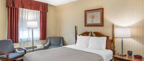 Quarto de hotel com uma cama grande, uma escrivaninha com cadeira, uma mesinha de cabeceira com abajur e um quadro na parede.