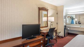 Une chambre d’hôtel avec un bureau en bois, une télévision, une chaise, un lit, un miroir et une petite kitchenette.