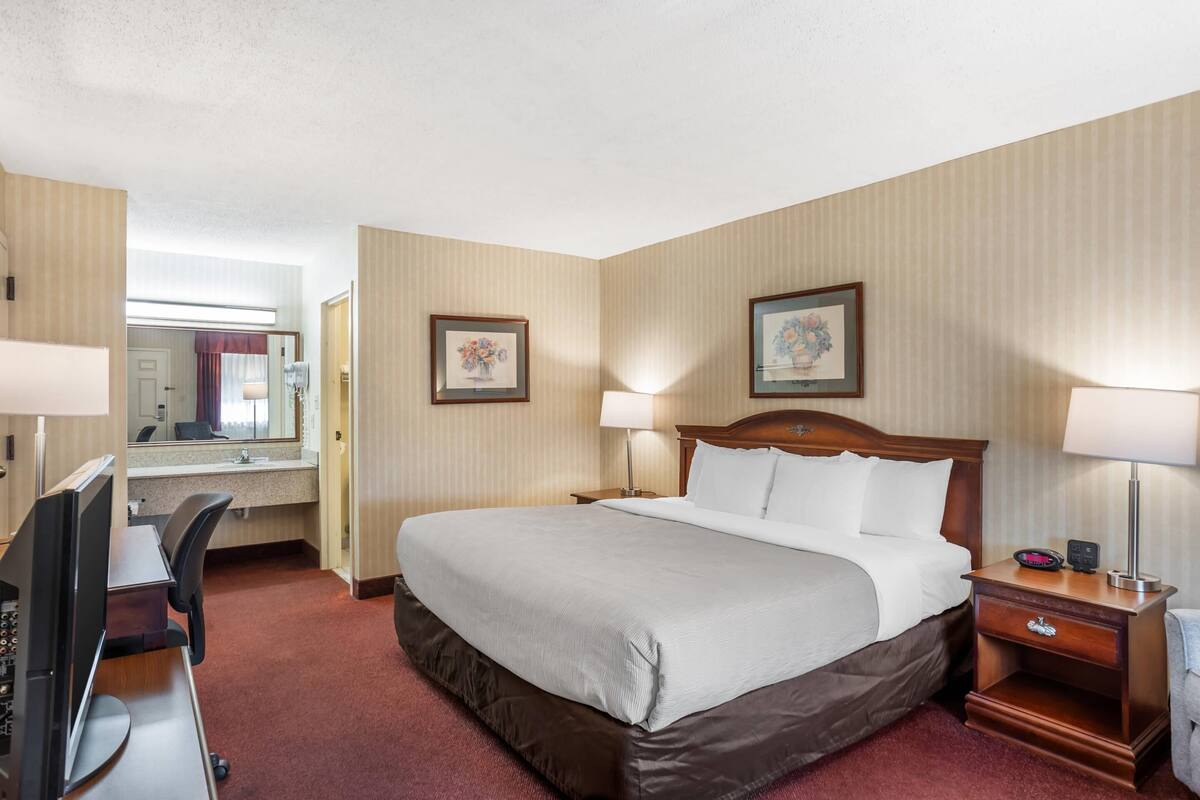 A hotel room with a large bed, a desk with a computer, a bedside table with a lamp, and a mirror.