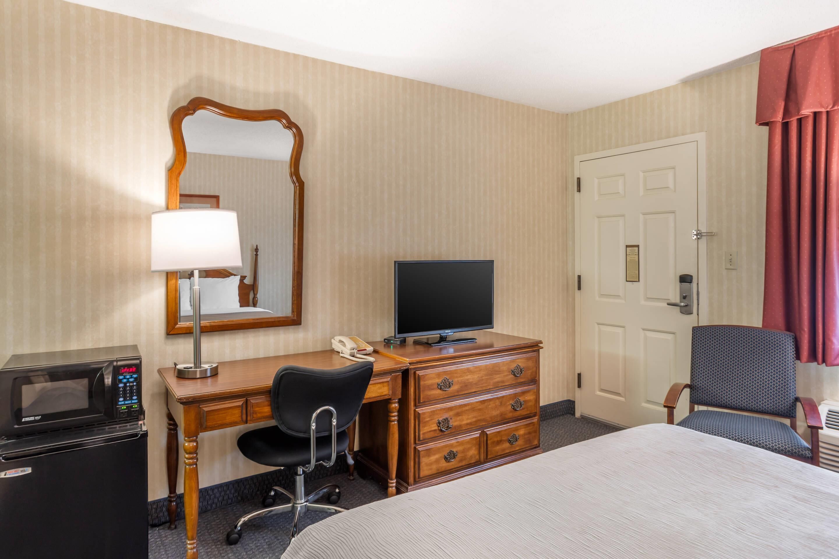 Habitación de hotel con una cama, un escritorio con computadora, un microondas, un teléfono, una silla y una puerta.