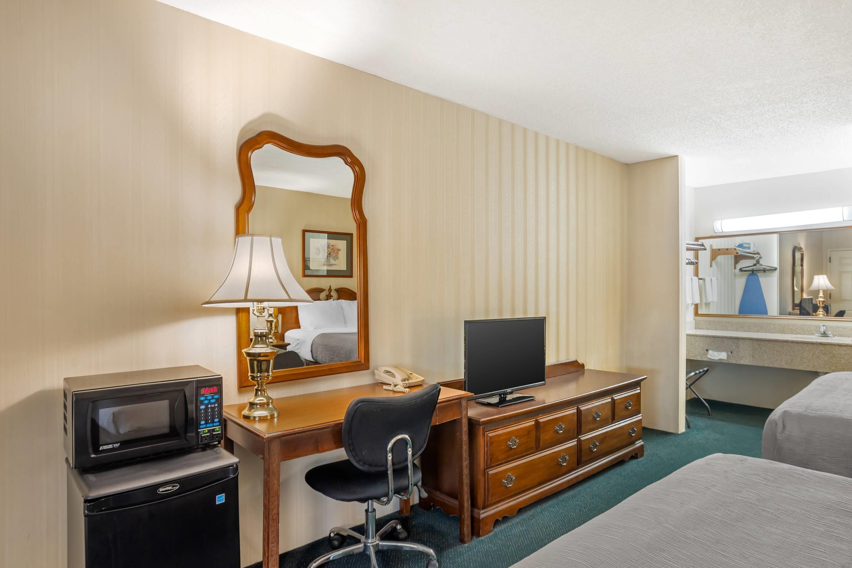 A hotel room with a desk, a chair, a dresser, a bed, a microwave, a telephone, and a television.