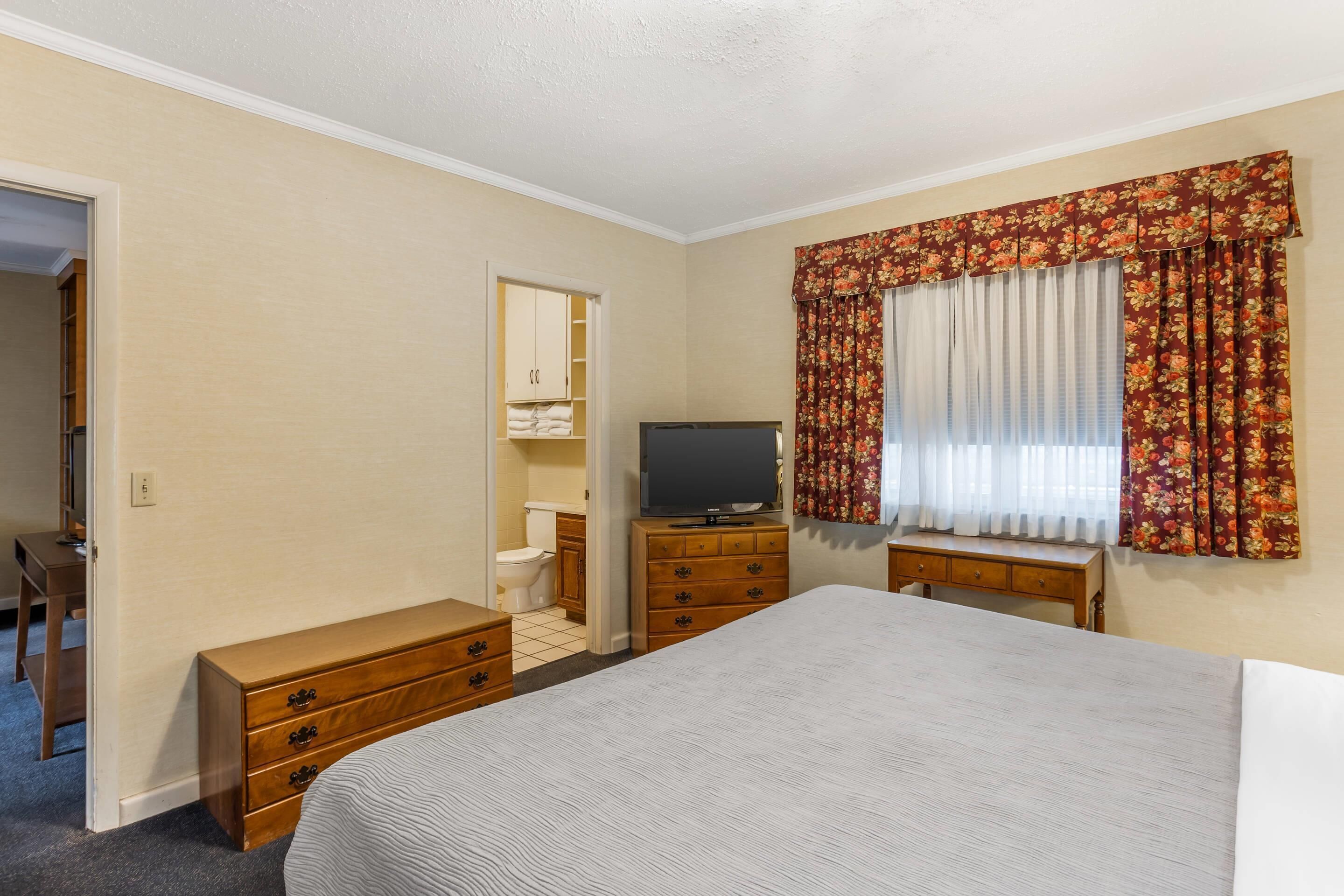 Un dormitorio con cama, cómoda, televisor, armario y ventana con cortinas.