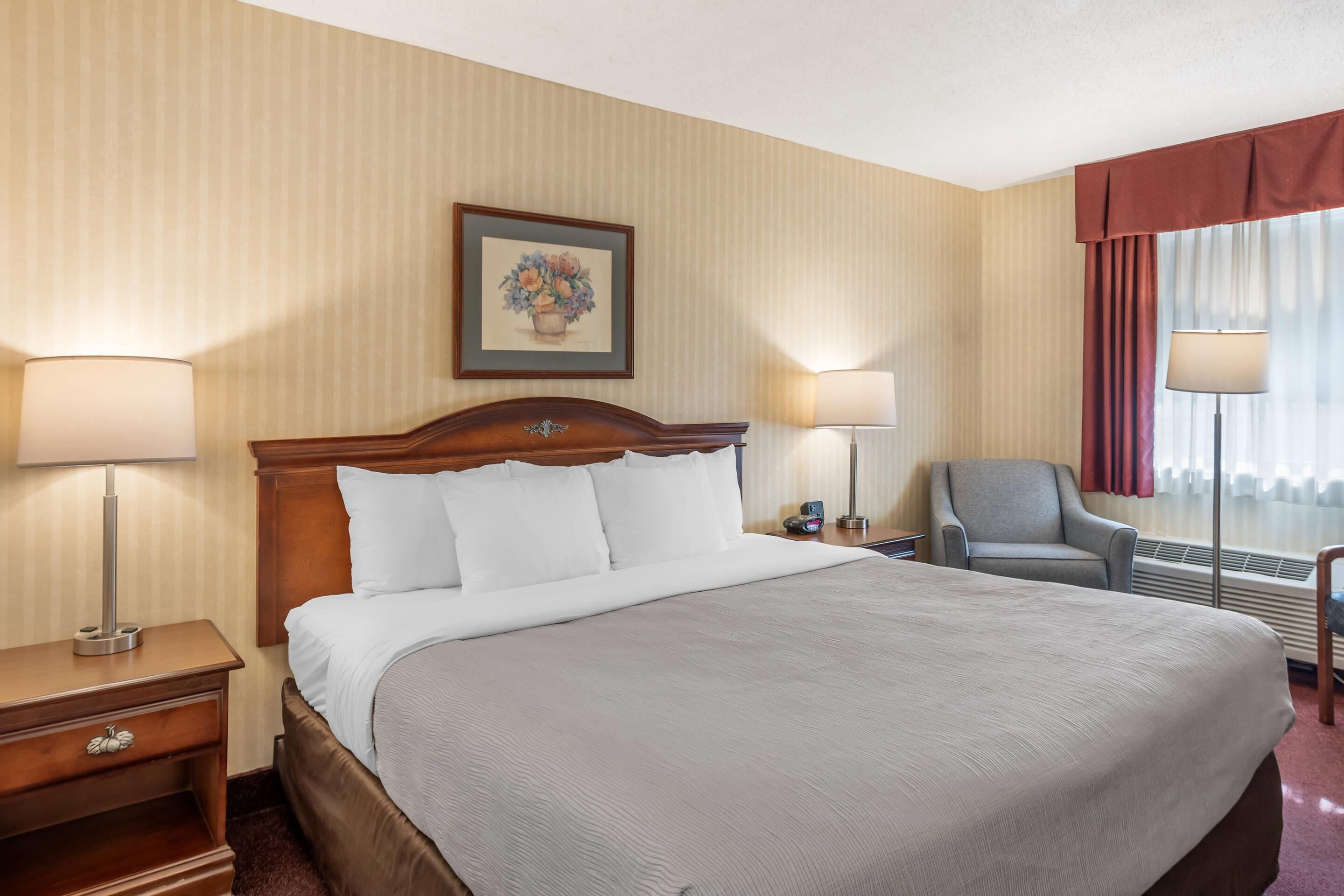 Une chambre d’hôtel avec un grand lit, deux tables de chevet avec des lampes, une chaise, un tableau encadré au mur et une fenêtre avec des rideaux.