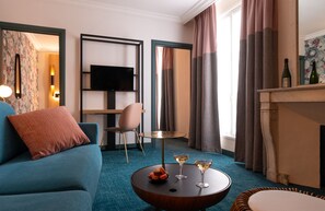 Una habitación de hotel moderna con un sofá azul, una mesa redonda de centro con bebidas y aperitivos, un televisor sobre un soporte y una chimenea con botellas.