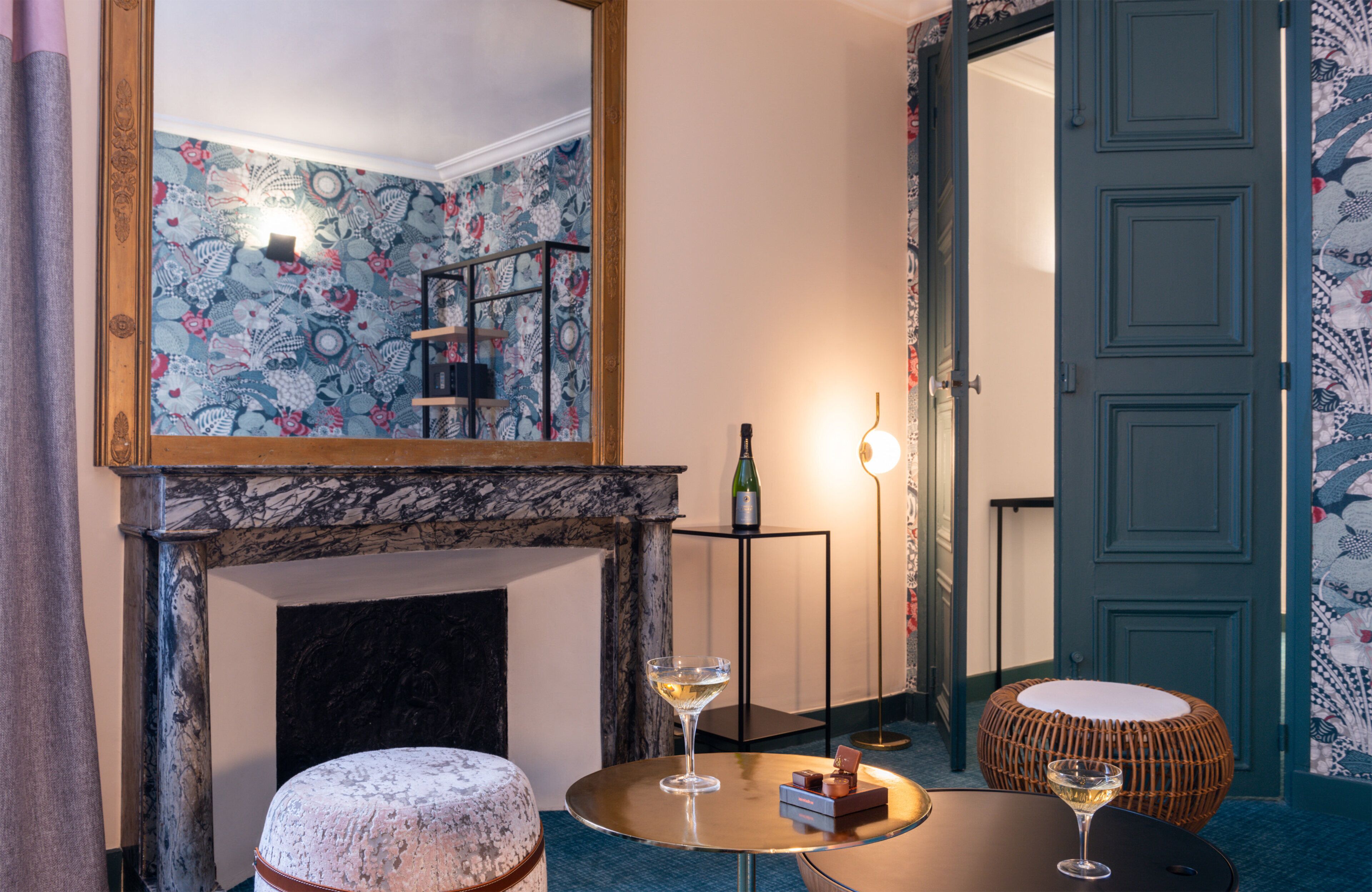 Una habitación con chimenea de mármol, una mesa redonda con dos copas de vino, un reposapiés de mimbre y un espejo que refleja la estancia.