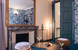 A room with a marble fireplace, a round table with two glasses of wine, a wicker ottoman, and a mirror reflecting the room. - Leonardo Boutique Hotel Paris Opera (Paris)