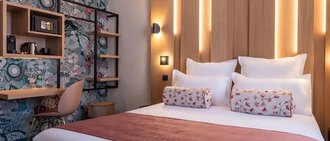 A hotel room with a large bed, a wooden headboard, a nightstand, a chair, and a shelf with books and a kettle.
