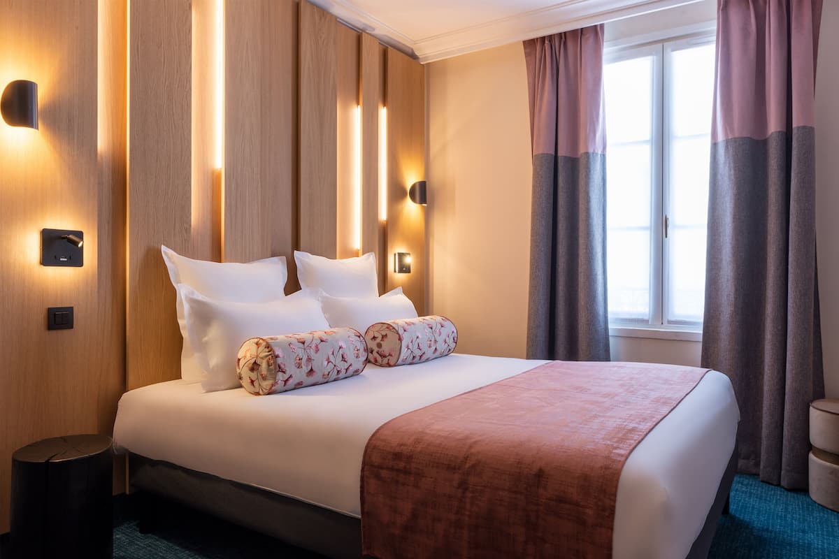 A neatly made bed with white linens and a brown throw, a wall-mounted light fixture, and a window with curtains.