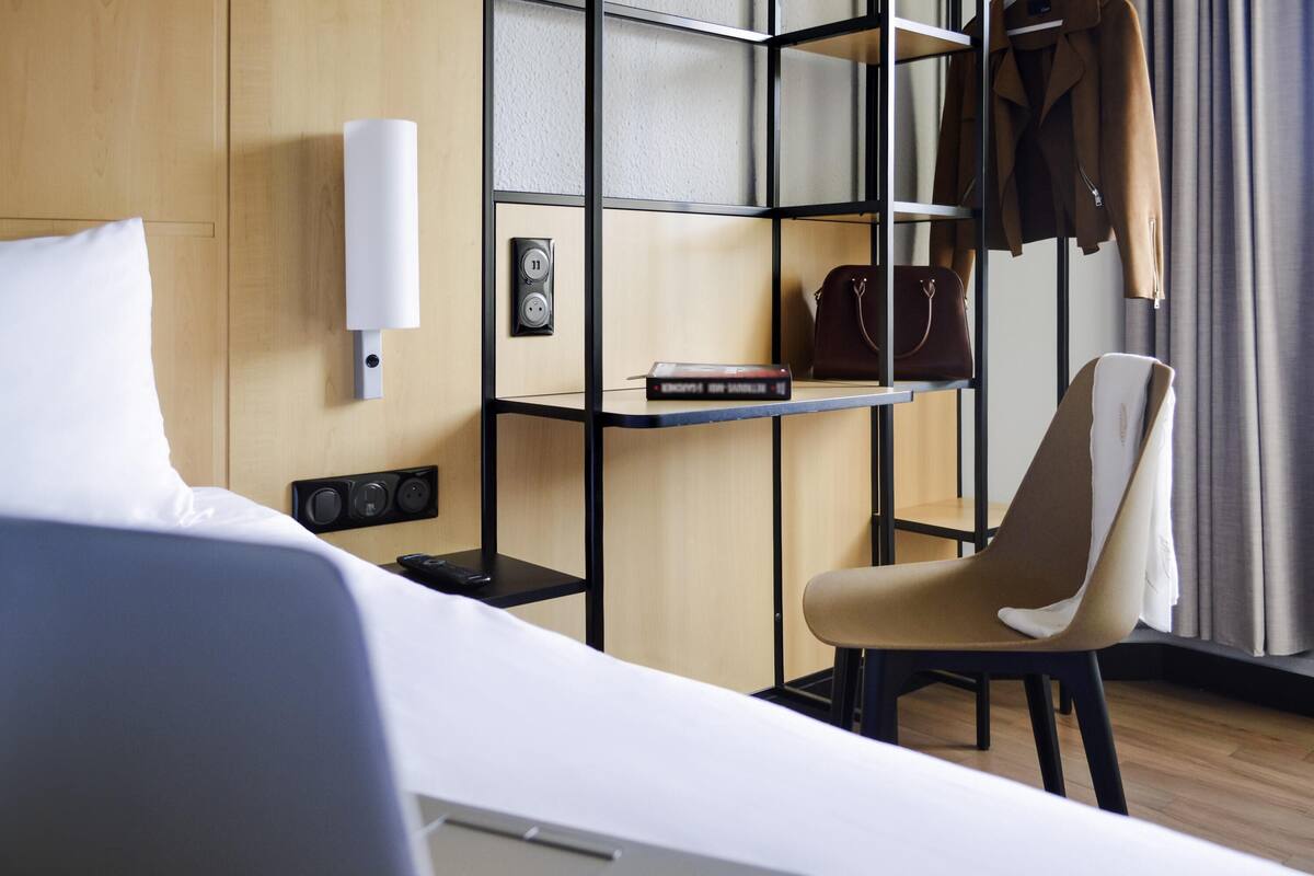 Ein Hotelzimmer mit einem Bett, einem Schreibtisch mit einem Laptop, einem Stuhl, einem Regal mit einem Buch und einer Handtasche sowie einer Wandsteckdose.