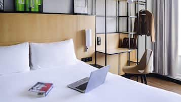 Habitación de hotel con cama, laptop, silla, perchero y repisa con bolso.