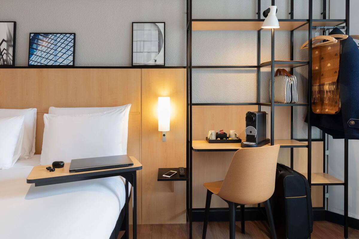Ein Hotelzimmer mit einem Bett, einem Schreibtisch mit einem Laptop, einem Sessel, einer Kaffeemaschine, einem Regal mit Taschen und einer Wandlampe.
