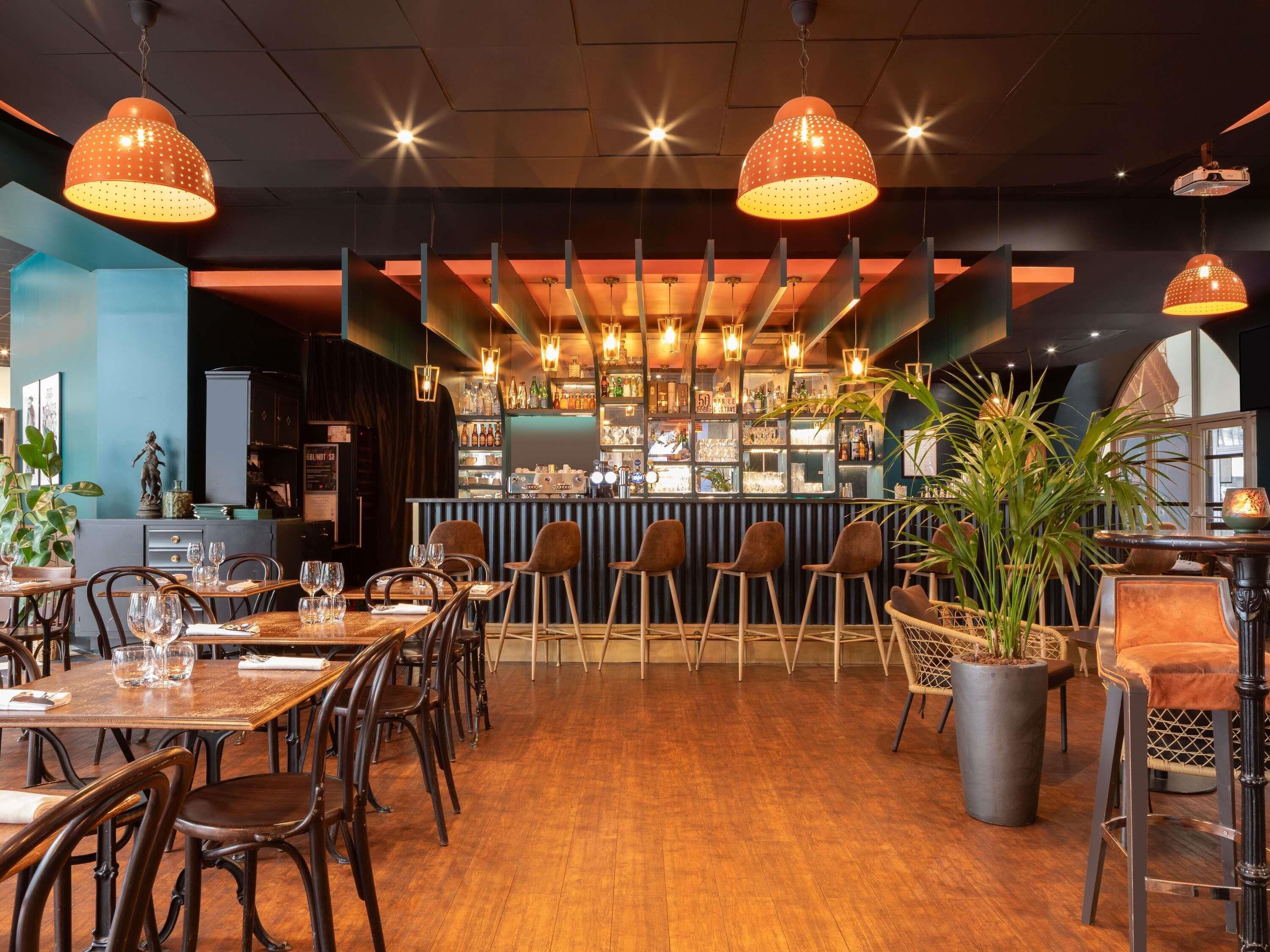 Een restaurantinterieur met houten tafels, stoelen, hangende lampen, een bar en potplanten.