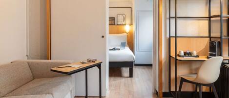 A hotel room with a sofa, a desk with a chair, a bed, and a shelf with a lamp.