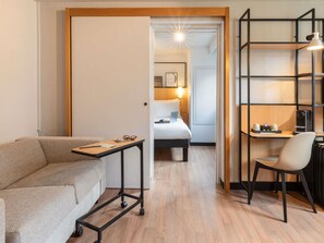 Quarto de hotel com sofá, mesa de trabalho com cadeira, cama e prateleira com luminária.