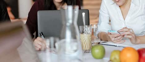 Dos personas sentadas a una mesa con una computadora portátil, un cuaderno y un teléfono inteligente. Hay vasos, una botella y frutas sobre la mesa.