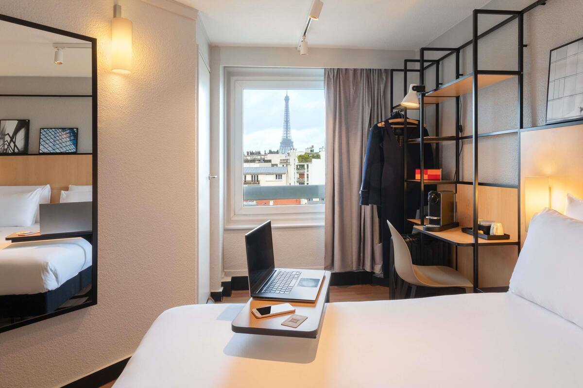 Ein Hotelzimmer mit Bett, Schreibtisch mit Laptop, Stuhl, Kleiderschrank und Blick auf den Eiffelturm.