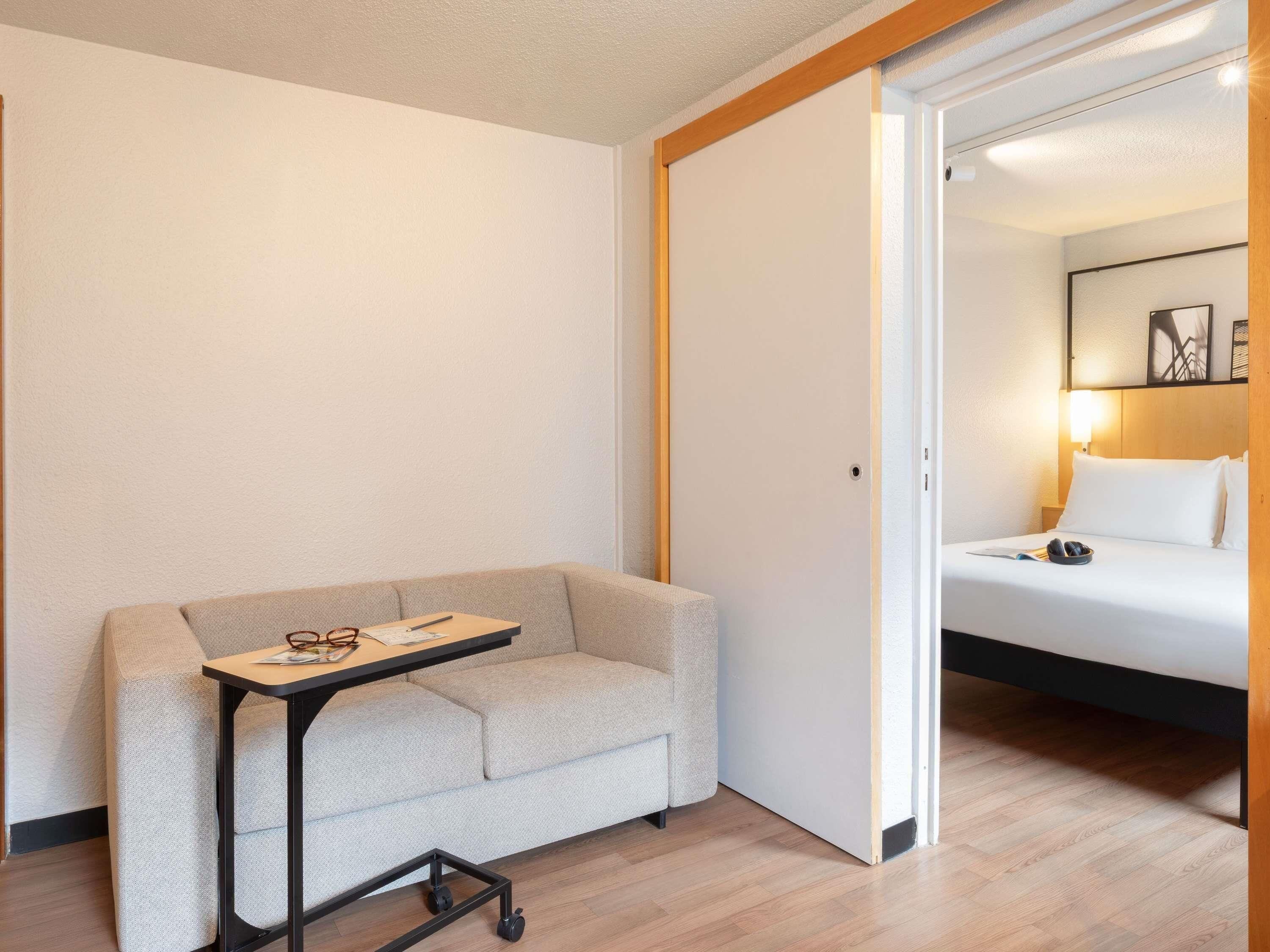 Ein Hotelzimmer mit einer Couch, einem kleinen Tisch mit Gläsern und einem Buch, sowie einem Bett mit Kissen und einer Lampe.