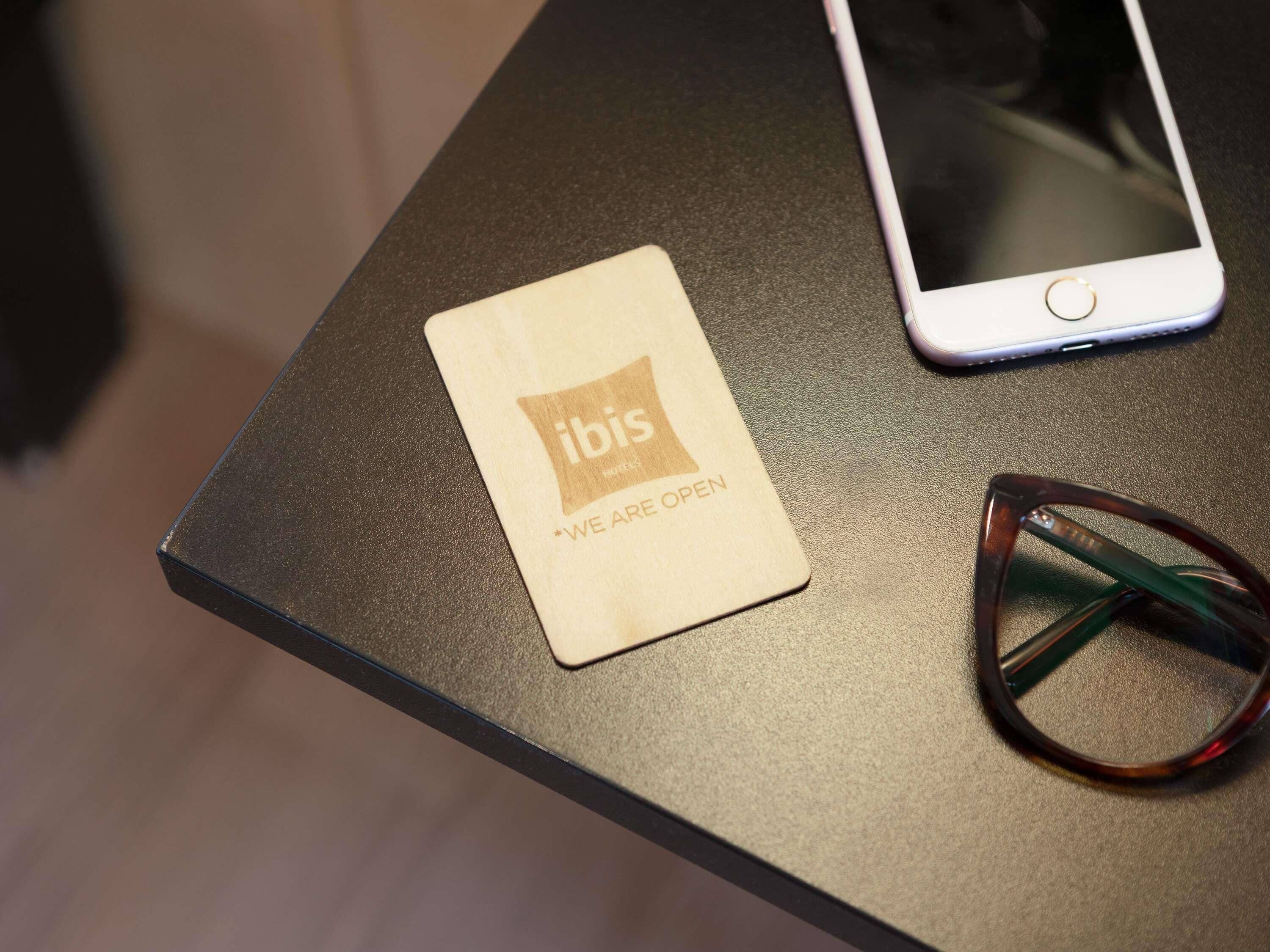A wooden card with 'ibis' and 'WE ARE OPEN' text, a smartphone, and a pair of sunglasses on a table.