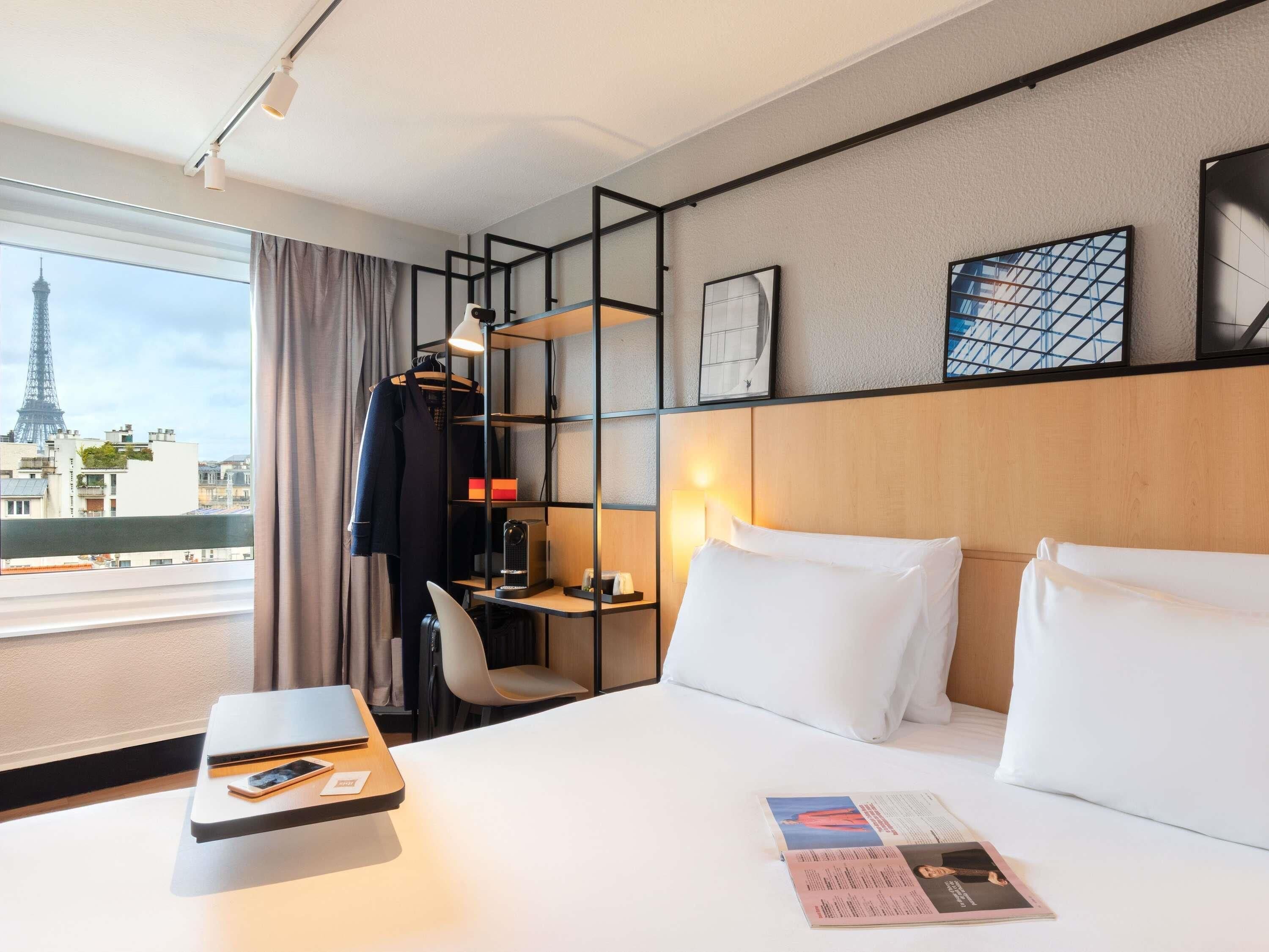 A hotel room with a bed, a desk, a chair, a wardrobe, and a view of the Eiffel Tower.