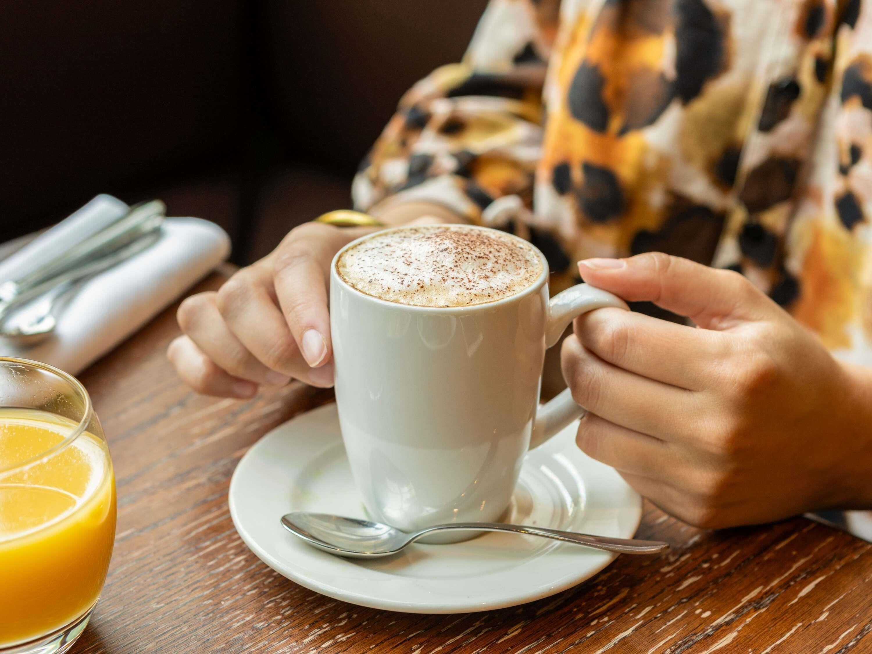 Una persona sosteniendo una taza blanca con un platito y una cuchara sobre una mesa de madera.