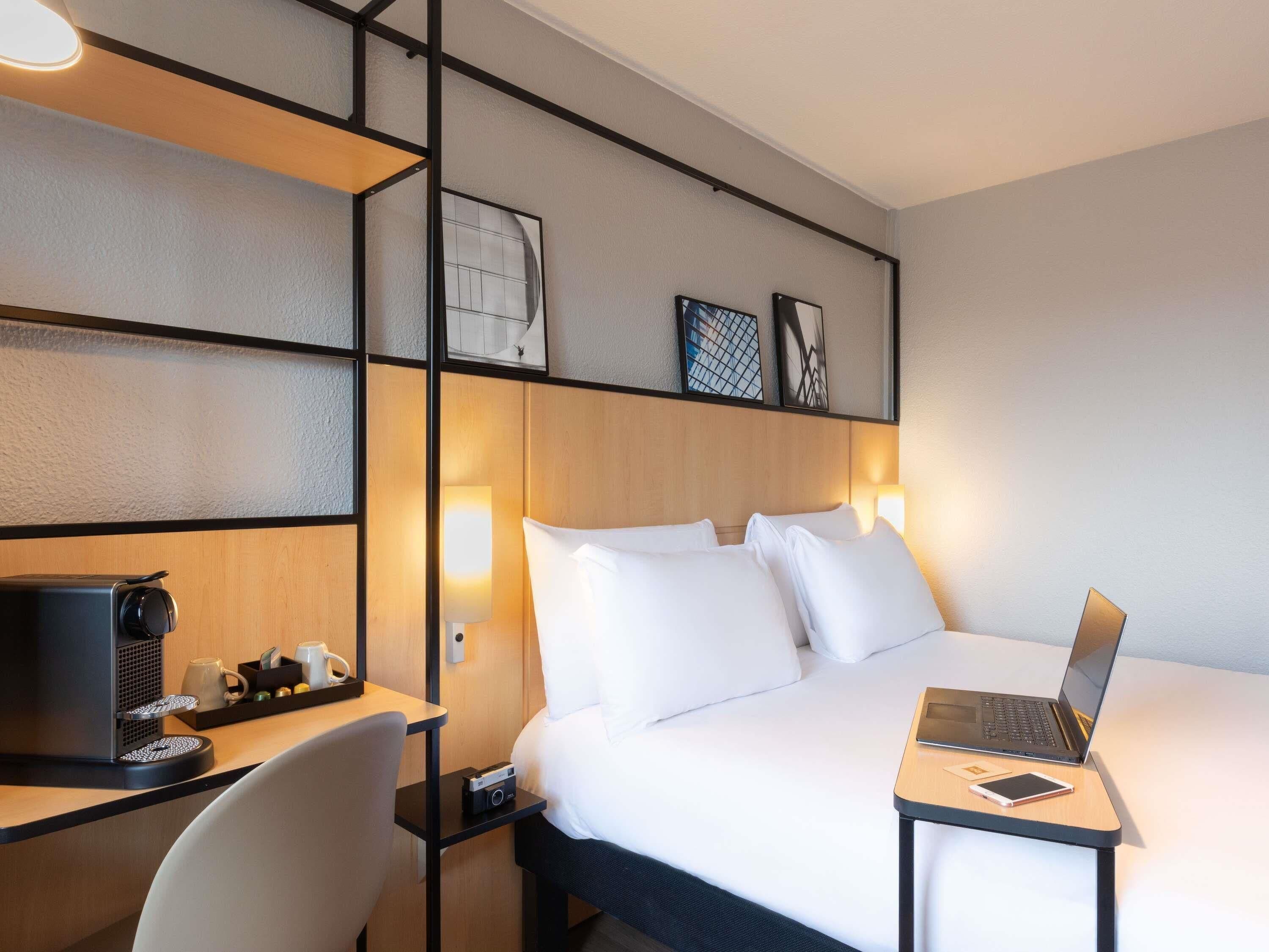 A hotel room with a bed, a desk with a laptop, a chair, a coffee machine, and a small table with a cup and a remote control.