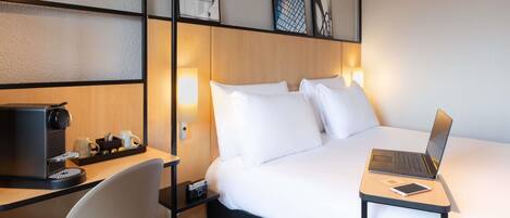 A hotel room with a bed, a desk with a laptop, a chair, a coffee machine, and a small table with a cup and a remote control.