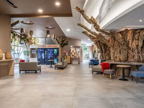 Een ruime lobby met een modern design, met daarin een grote boomsculptuur, zitgedeeltes, automaten en een bordje met 'IBIS SNACKING'.