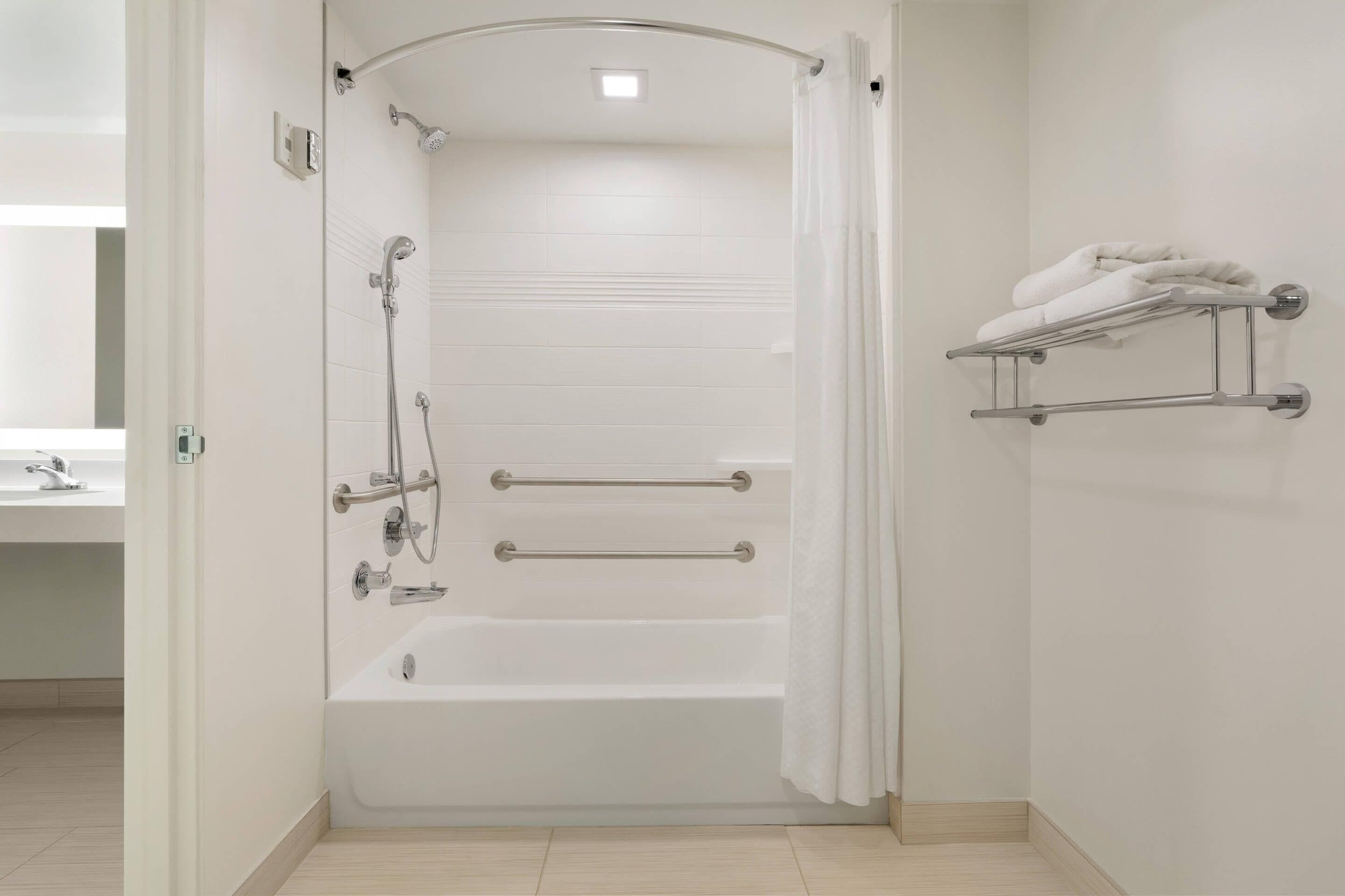 Un baño moderno con cabina de ducha, barras de apoyo, toallero y suelo de baldosas blancas.