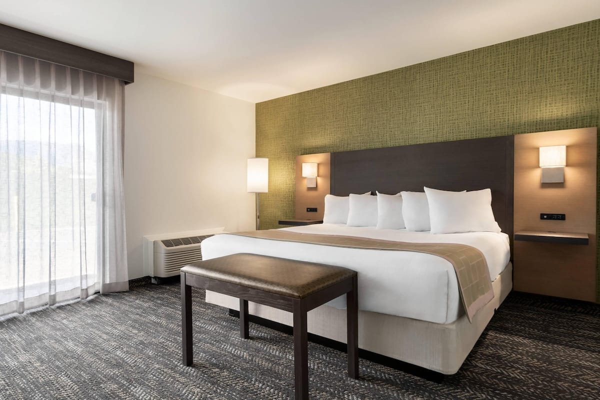 A hotel room with a large bed, two bedside tables, a bench, a window with sheer curtains, and a wall-mounted air conditioner.