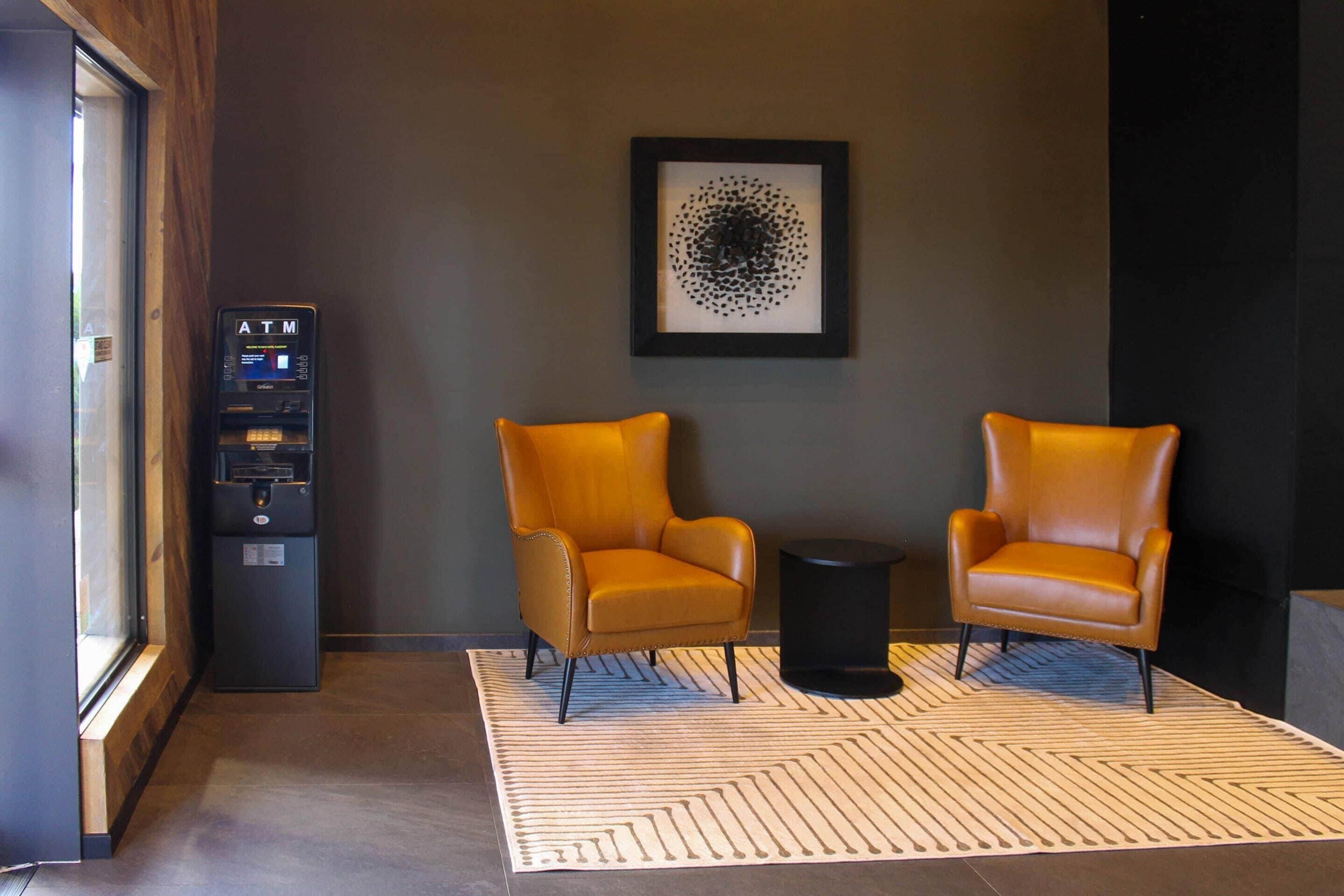 A lobby with two leather armchairs, a small table, an ATM, and a framed abstract artwork on the wall.