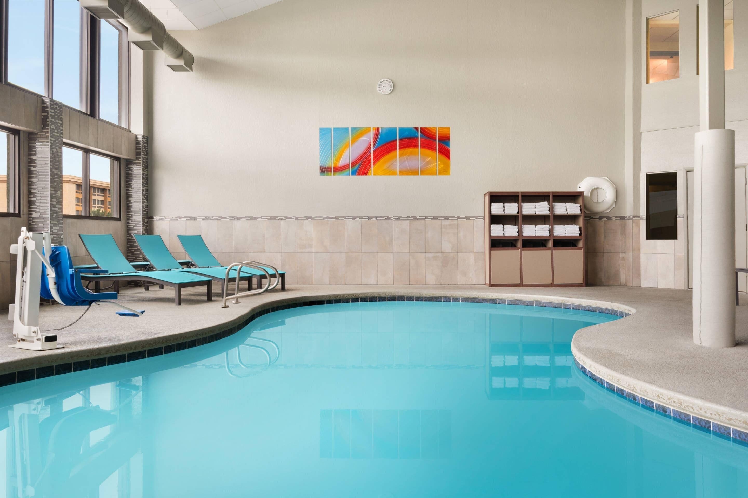 An indoor swimming pool with blue lounge chairs, a wall-mounted clock, and a colorful abstract painting.