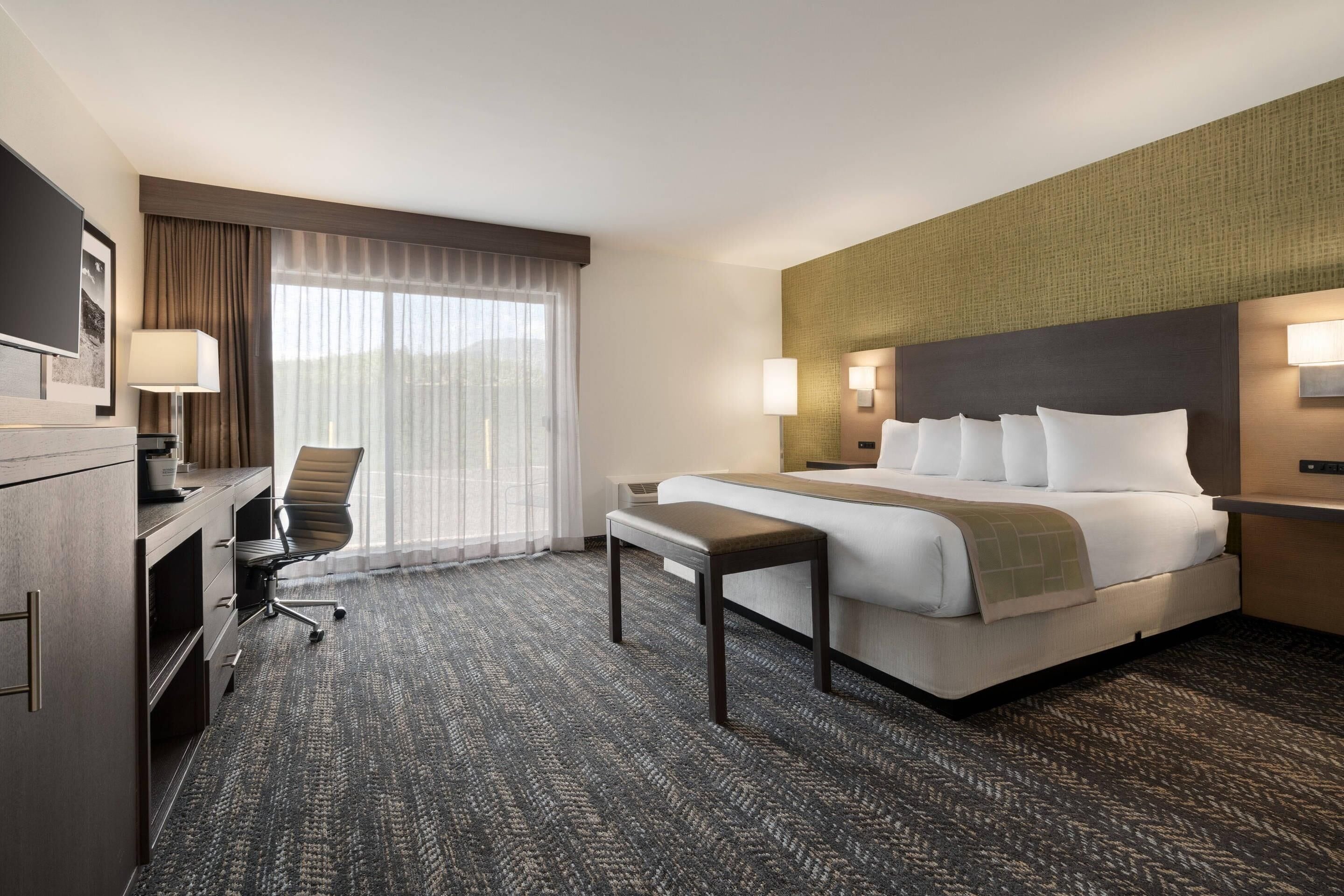 Quarto de hotel com cama, escrivaninha, cadeira e uma grande janela com cortinas.