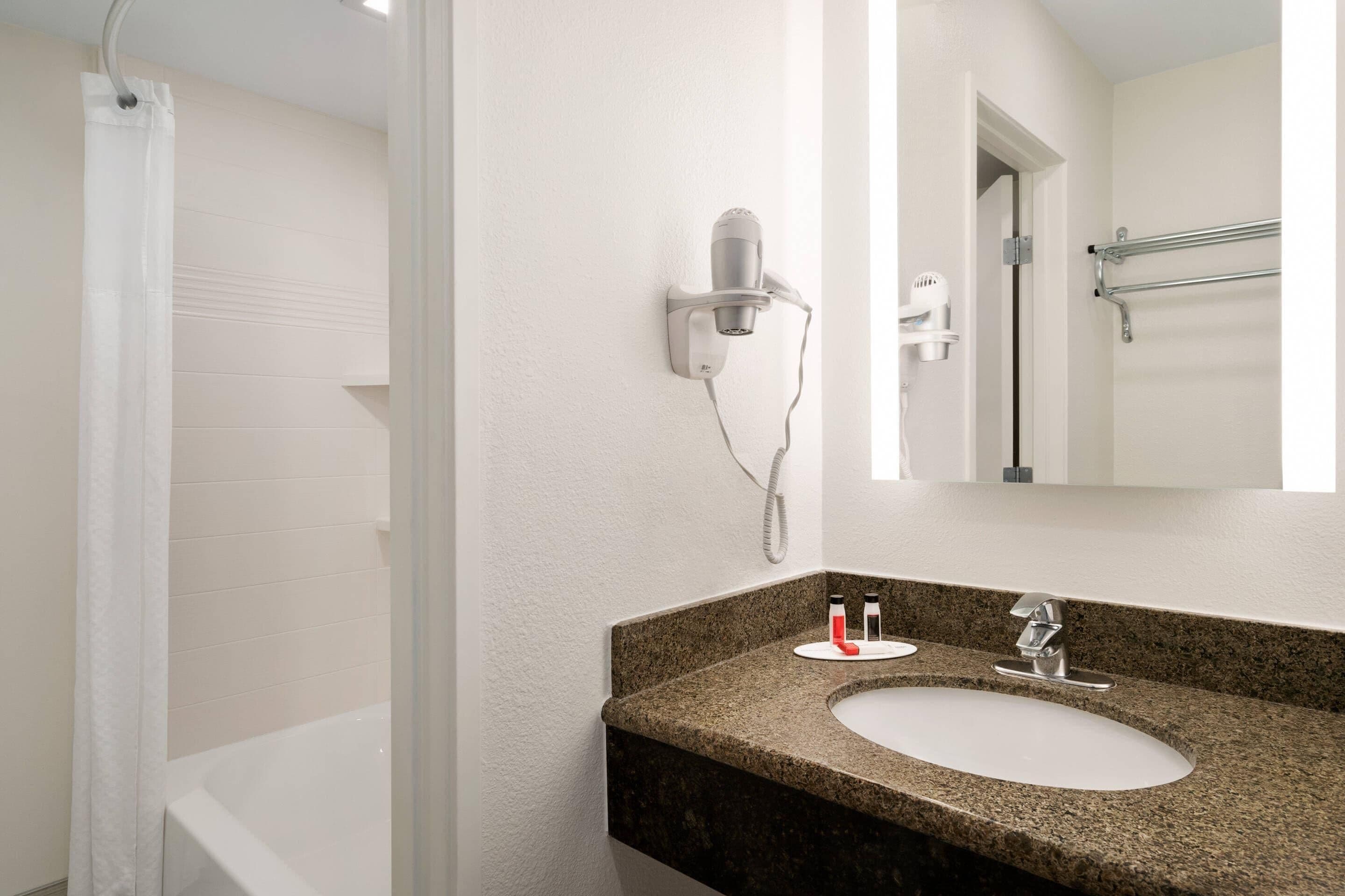 Une salle de bain avec une baignoire blanche, un lavabo avec un plan de travail en granit, un sèche-cheveux fixé au mur et un porte-serviettes.