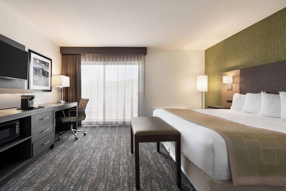 Quarto de hotel com cama, escrivaninha com cadeira, televisão e janela com cortinas.
