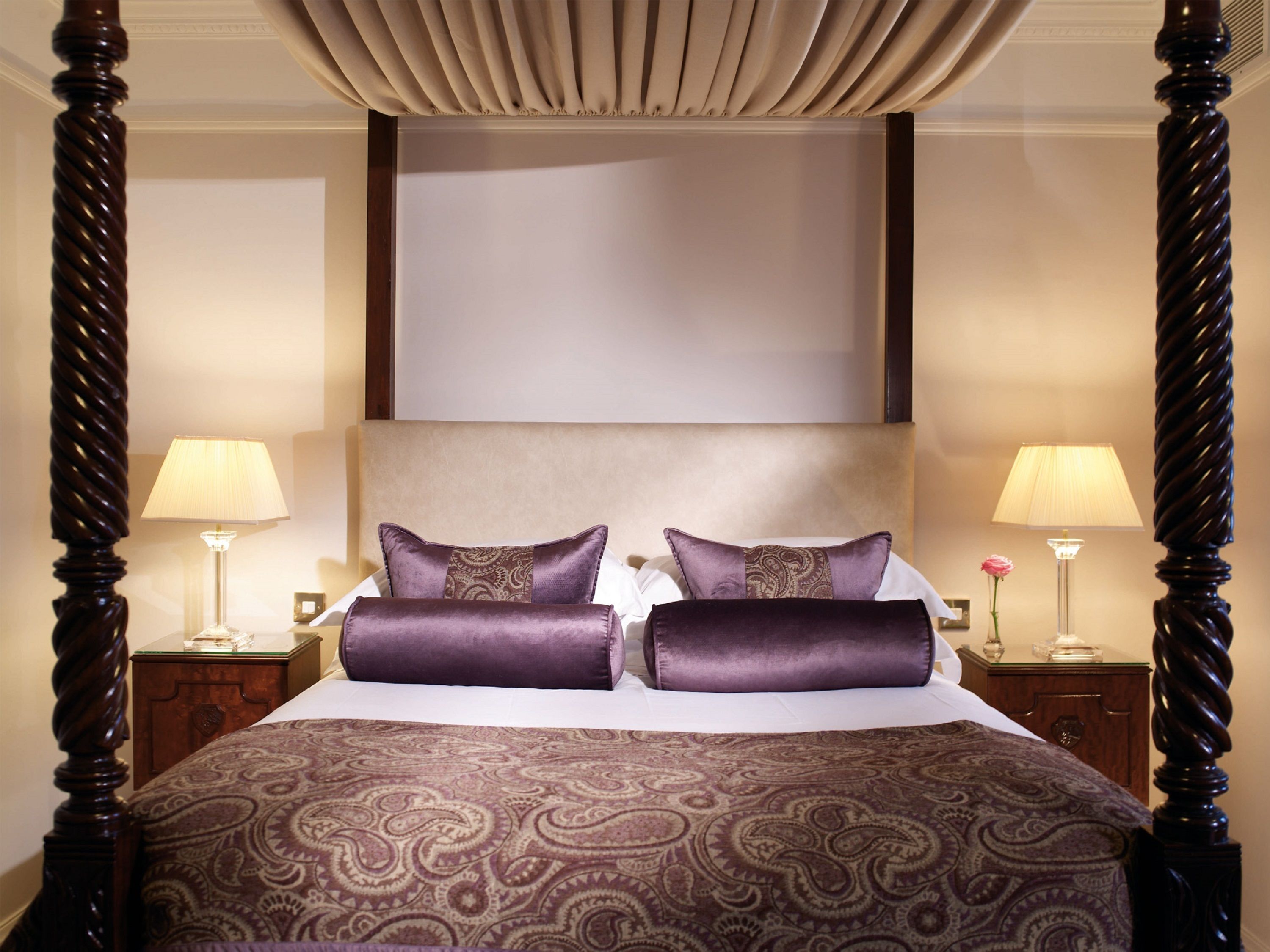 A four-poster bed with a patterned bedspread, two matching pillows, and a headboard. There are two bedside tables with lamps and a vase of flowers.