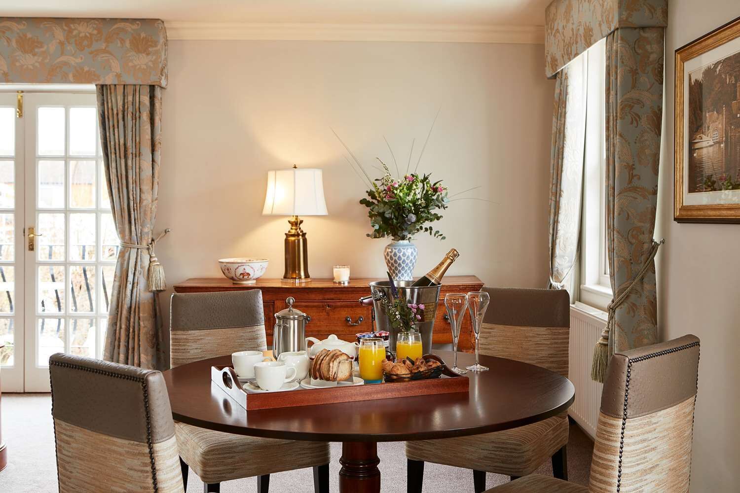 A dining area with a round table, chairs, a lamp, a vase with flowers, a bottle of champagne, and breakfast items.