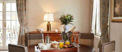 A dining area with a round table, chairs, a lamp, a vase with flowers, a bottle of champagne, and breakfast items.